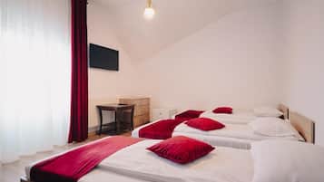 Quarto de hotel com três camas, cada uma com lençóis brancos e almofadas vermelhas. Há uma secretária de madeira, uma cadeira e uma televisão de tela plana na parede.
