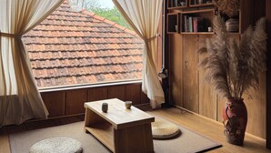 Une pièce de style japonais traditionnel, avec un sol recouvert de tatamis, une petite table en bois, une bibliothèque et une vue sur un toit en tuiles à l’extérieur.