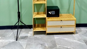 Une pièce avec un mur vert, un lampadaire noir sur trépied, un meuble de rangement en bois à plusieurs niveaux sur lequel se trouve un appareil électronique noir, et un porte-manteau en bois.