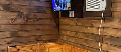 Un interior de madera con una gran mesa de madera y bancos, un televisor montado en la pared y un equipo de aire acondicionado blanco.