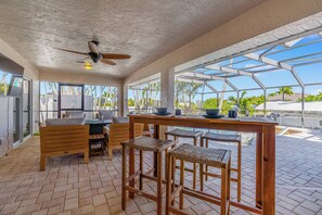 Un vaste espace extérieur aménagé pour les repas, avec des meubles en bois, un ventilateur de plafond et de grandes fenêtres offrant une vue sur les palmiers et un ciel dégagé.