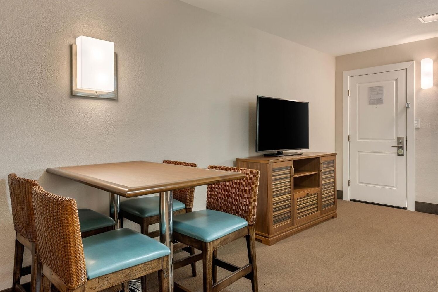 Una habitación de hotel con una mesa de comedor, cuatro sillas, un televisor sobre un mueble de madera y una puerta blanca.