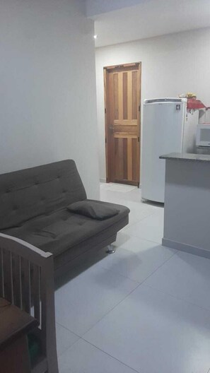A living room with a sofa and a small table, a wooden door, and a kitchen area with a refrigerator. - Charming 1-bedroom apartment in Armação dos Búzios perfect for relaxing (Armação dos Búzios)