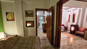 Ein Zimmer mit einem Bett, einem Holzschrank, einem Spiegel und einem kleinen Tisch mit einer Lampe. Ein Flur führt in ein weiteres Zimmer mit einem roten Sessel und einem Heizgerät.