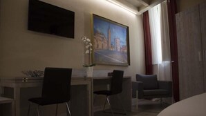 A hotel room with a desk, two chairs, a bed, a painting of a cityscape, and a large window with curtains. - Palazzo Domanto Parma Apartments (Parma)