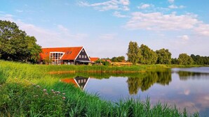 Une scène paisible au bord d’un lac, avec une maison traditionnelle dotée d’un toit rouge et de grandes fenêtres, entourée d’une végétation luxuriante et d’arbres.