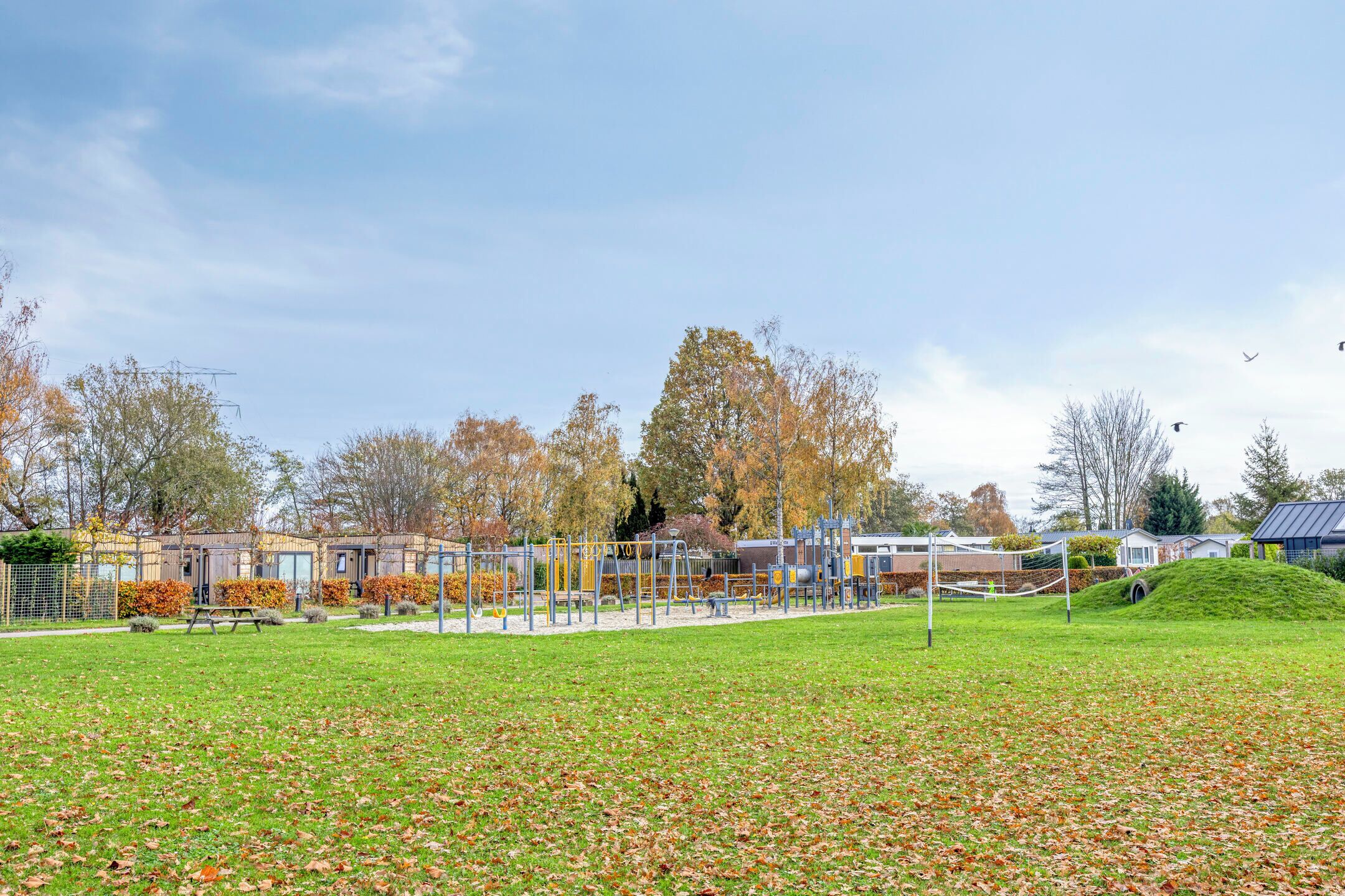 Ein Spielplatz mit Schaukeln und Klettergerüsten, umgeben von Bäumen und einem Gebäude mit einem geneigten Dach.