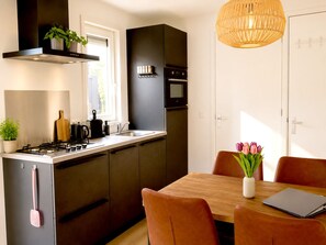 Een moderne keuken met een gasfornuis, oven en magnetron, een eethoek met tafel en stoelen, en een hanglamp.
