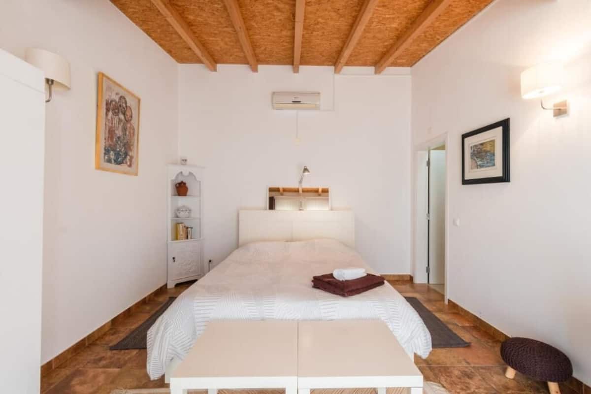 Quarto com cama, mesa de cabeceira, luminária de parede e uma pequena prateleira com objetos decorativos.