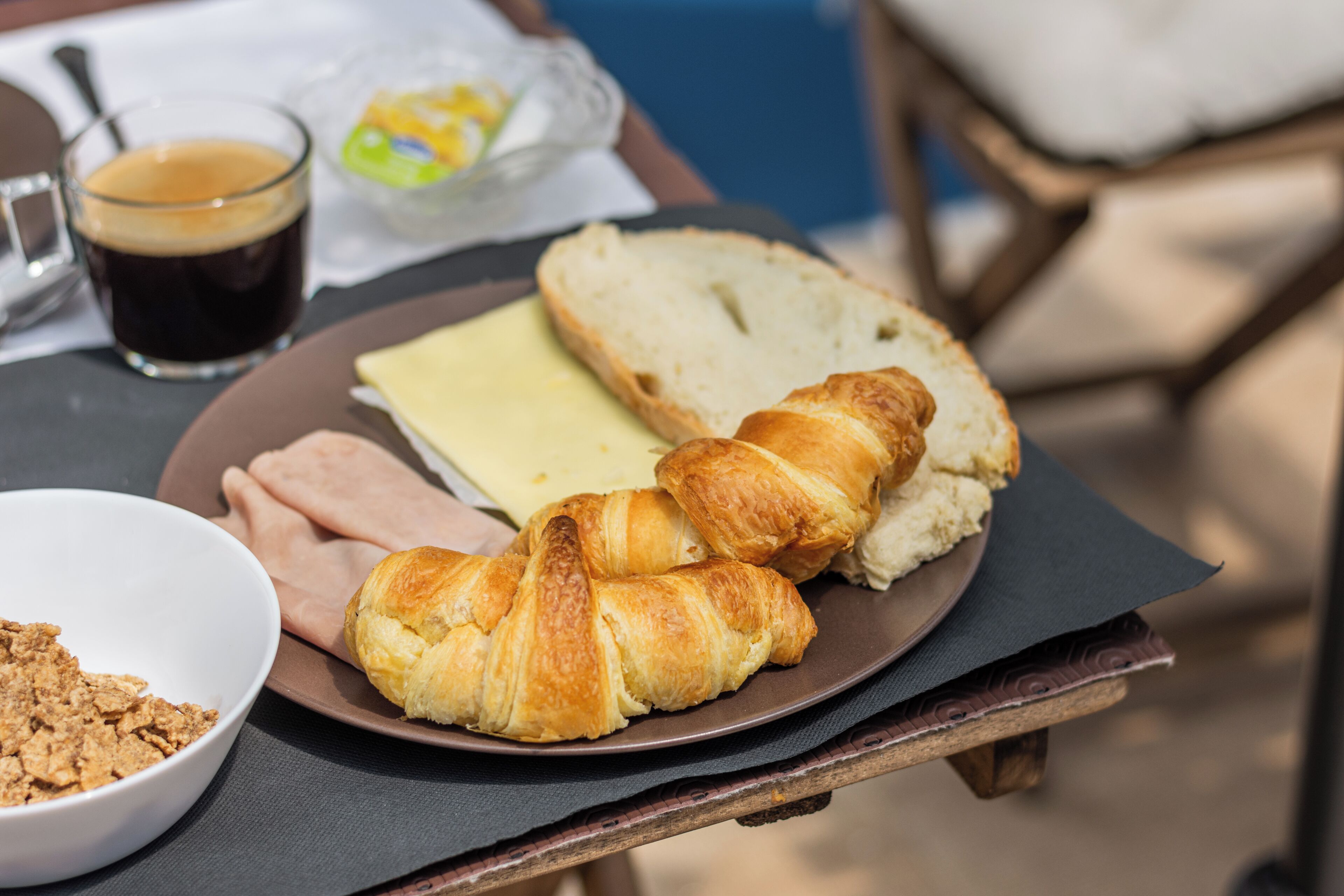 A breakfast plate with croissants, cheese, and ham, a glass of coffee, and a bowl of cereal on a table.