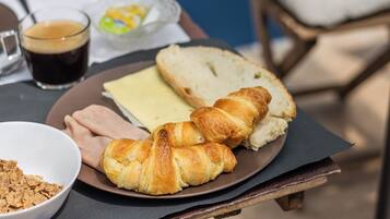 A breakfast plate with croissants, cheese, and ham, a glass of coffee, and a bowl of cereal on a table.
