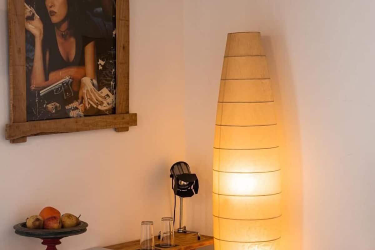 Um quarto com um quadro emoldurado em madeira, um candeeiro alto, uma fruteira e uma cadeira.