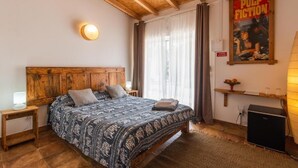 Une chambre avec une tête de lit en bois, un lit avec une literie à motifs, une table de chevet, une lampe fixée au mur, une affiche, une fenêtre avec des rideaux et une petite étagère avec quelques objets.