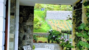 A covered patio with a stone wall, a stone floor, and a view of a garden with plants and flowers. - Pensive Villa - Nuwara Eliya (Nuwara Eliya)