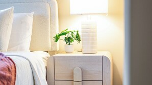 A neatly made bed with white bedding and a pink throw, a bedside table with a lamp and a potted plant, and wooden flooring. - 4BR LU Getaway! 4 Min to LU • Hot Tub • Game Room (Lynchburg)