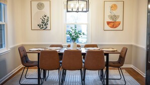 A dining room with a rectangular table, six brown chairs, a vase with green plants, and two framed abstract artworks on the walls. - 4BR LU Getaway! 4 Min to LU • Hot Tub • Game Room (Lynchburg)