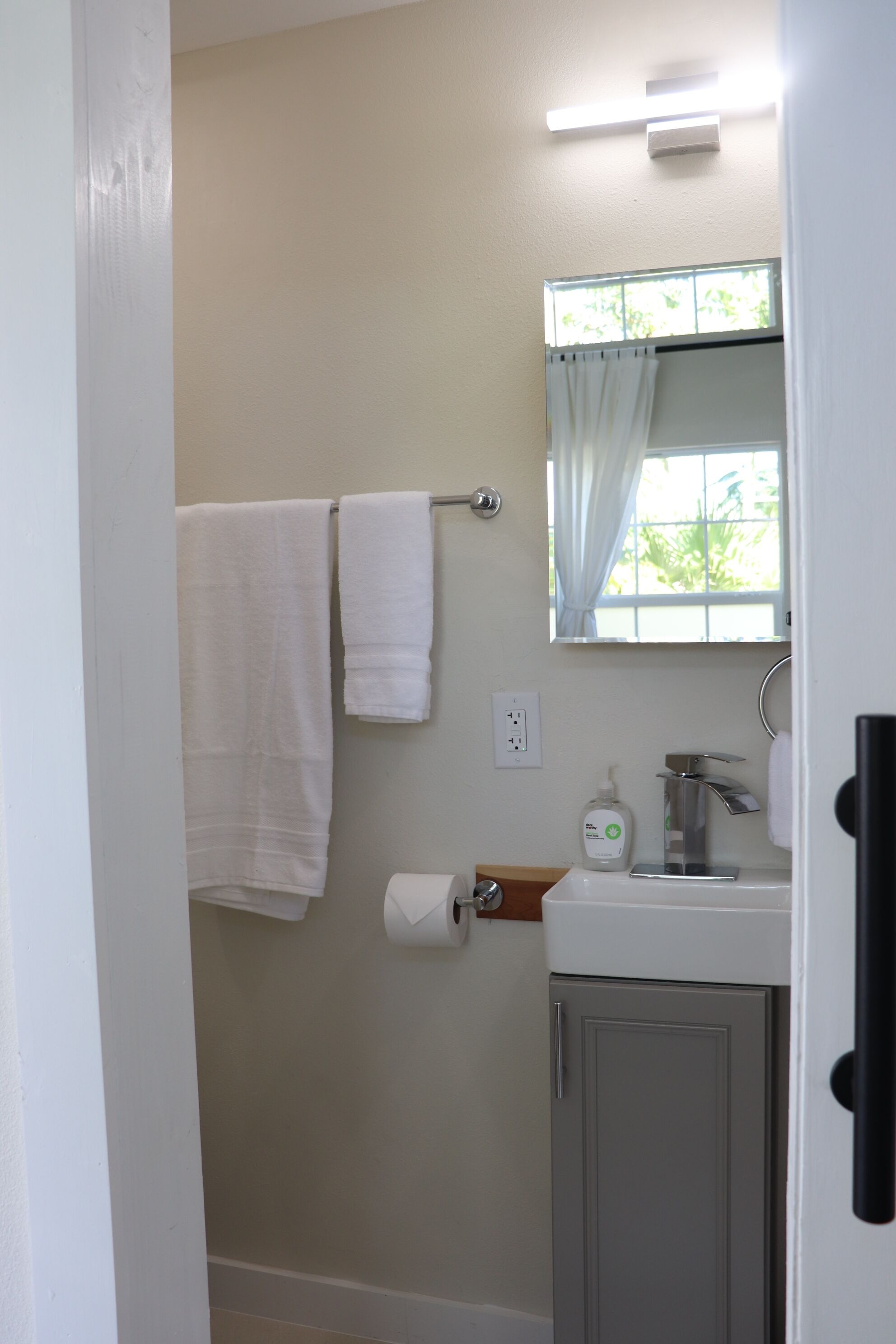 Une salle de bain avec un lavabo blanc, un miroir, un porte-serviettes avec deux serviettes, une fenêtre avec un rideau et un porte-rouleau de papier toilette.