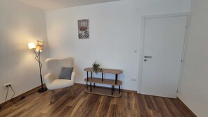 Una habitación con una silla blanca, una lámpara de pie, una pequeña mesa de madera y una puerta blanca.