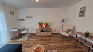 Una sala de estar moderna con suelo de madera, un televisor sobre un mueble de madera, un sofá blanco, una mesita con tazas y un escritorio con una silla.
