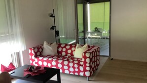En stue med en sofa med prikker, et lille bord og en stol. Der er en skydedør, der fører ud til en balkon med havemøbler.