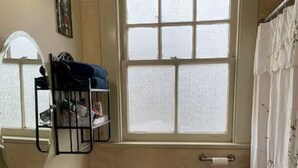 Une salle de bain avec un lavabo, des toilettes et une baignoire, tous de couleur blanche. Il y a une fenêtre au-dessus des toilettes et une étagère avec des articles de toilette.