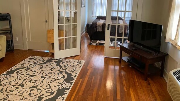 Une pièce avec un plancher en bois, une télévision, un lit recouvert d’une couverture sombre et une grande fenêtre avec des rideaux.