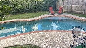 Une piscine bordée de briques, deux chaises rouges et un espace recouvert de gazon synthétique vert.