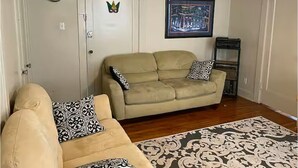 Un salon avec un canapé beige, des coussins à motifs, un tapis à motifs, une horloge, un tableau encadré et une porte agrémentée d’un élément décoratif.