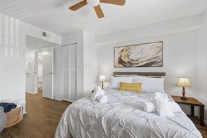 Ein Hotelzimmer mit einem Bett, zwei Nachttischlampen, einem Deckenventilator, einem Gemälde an der Wand und einer Tür zu einem Badezimmer.