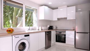 A modern kitchen with white cabinets, stainless steel appliances, a washing machine, and a window with a view of greenery.