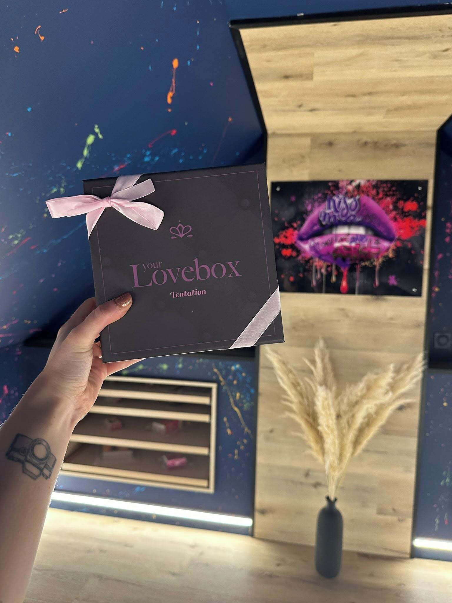 Iemand houdt een donkerbruine doos met lint vast, met de tekst 'Lovebox Tentation', in een kamer met een houten vloer, een muur met een kleurrijk abstract schilderij en een vaas met gedroogde planten.