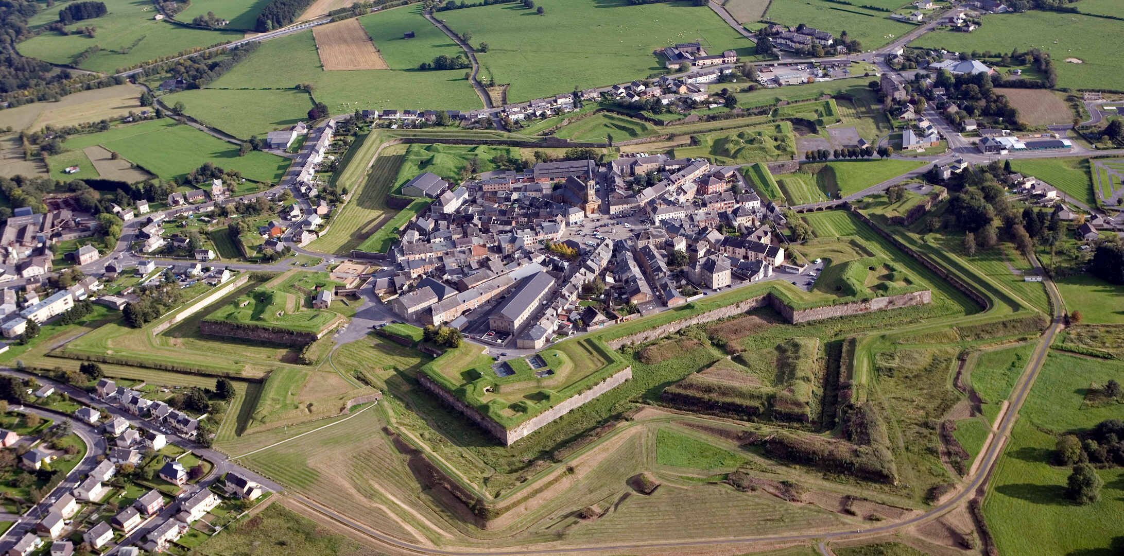 Luchtbeeld van een historische stad met een ommuurd kasteel, omgeven door groene velden en woonwijken.