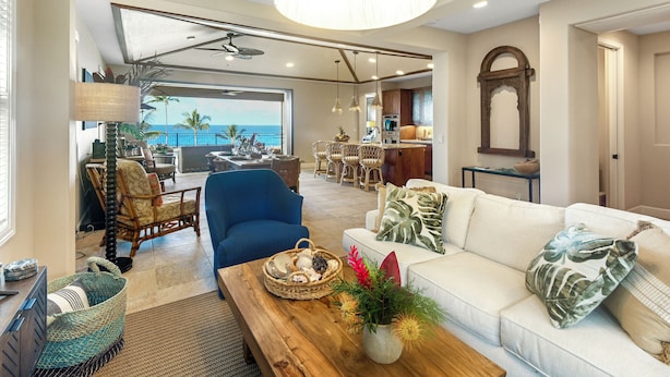A spacious living room with a white sofa, blue armchair, and a wooden coffee table. A large window offers a view of the ocean. - Blue Lani at Holua Kai (Kailua-Kona)