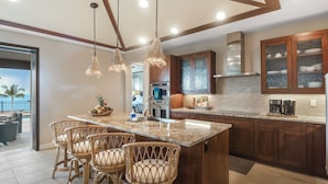 A kitchen with a large island, bar stools, wooden cabinets, and a view to the outside. - Blue Lani at Holua Kai (Kailua-Kona)
