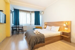 Quarto de hotel com cama de madeira, uma mesa com cadeiras, televisão e janela com cortinas.