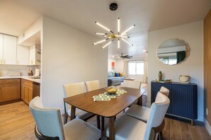 A modern dining area with a wooden table, six chairs, and a decorative ceiling light. A kitchen with wooden cabinets and a dishwasher is visible in the background. - Arcadia 2BR Condo Near Camelback and Old Town (Phoenix)