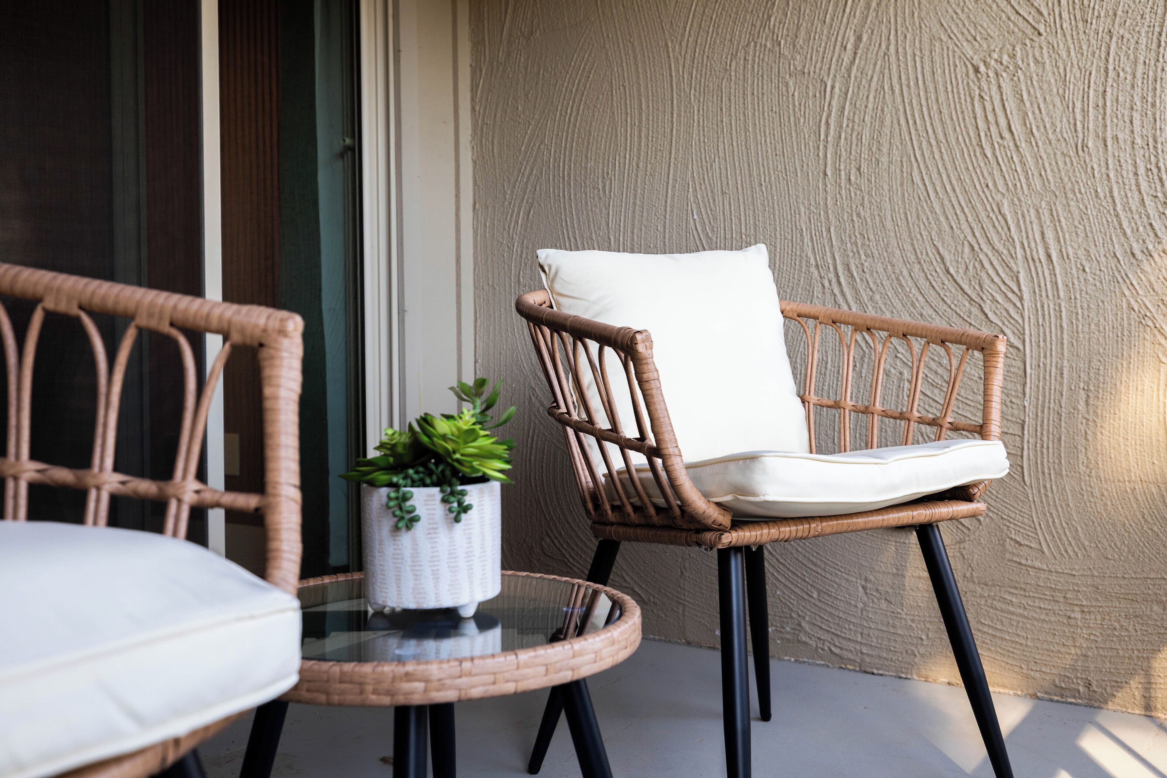 Une chaise en osier avec un coussin blanc et une petite plante en pot, posée sur une table en verre.
