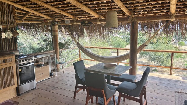 Um deck de madeira com mesa e cadeiras, uma rede e uma área de churrasco com forno e panelas.
