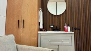 Quarto com guarda-roupa de madeira, espelho redondo, pia branca e sofá bege.