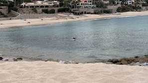 A beach with clear water, a person swimming, and buildings in the background. - Cozy one bedroom bungalow in beautiful San Jose del Cabo with AC Near the beach! (San Jose del cabo)
