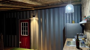 A compact, rustic bathroom with a red door, a wooden ceiling, and a metal wall. A hanging light fixture and a mirror are visible. - Cozy one bedroom bungalow in beautiful San Jose del Cabo with AC Near the beach! (San Jose del cabo)
