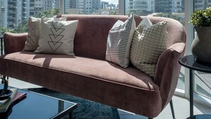 Una sala de estar moderna con un sofá rosa, una mesa de centro de vidrio y vistas a edificios altos a través de amplios ventanales.