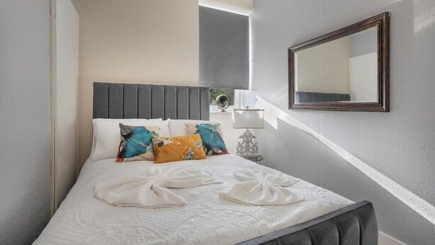 A neatly made bed with a grey headboard, white bedding, and colorful pillows. A small table with a lamp and a globe is beside the bed. - Holburn Lodge by Duthie Park Work Ready Fast Wi-fi (Aberdeen)