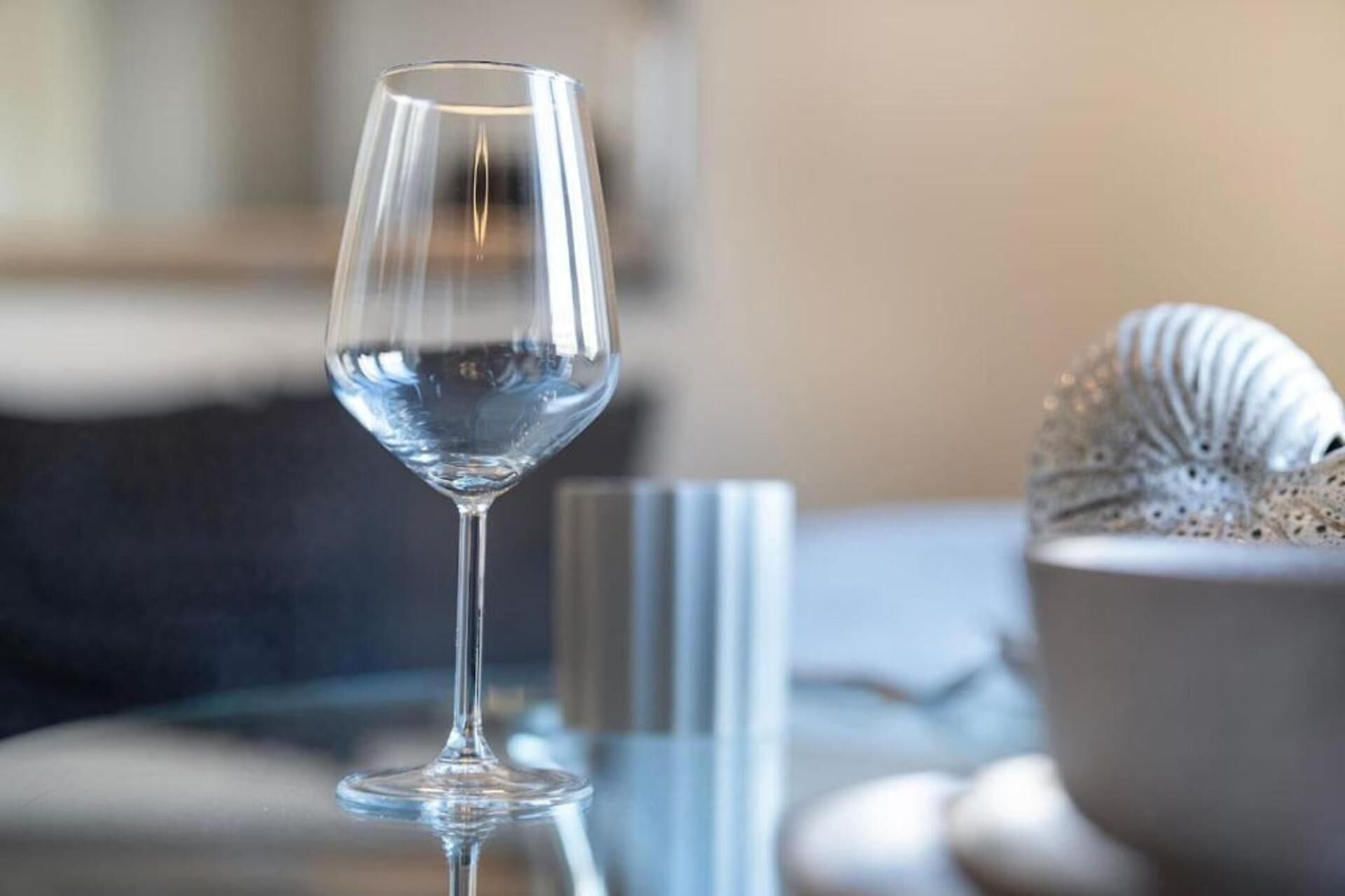 A clear glass wine glass on a reflective surface, possibly a table, with a blurred background suggesting a domestic setting.