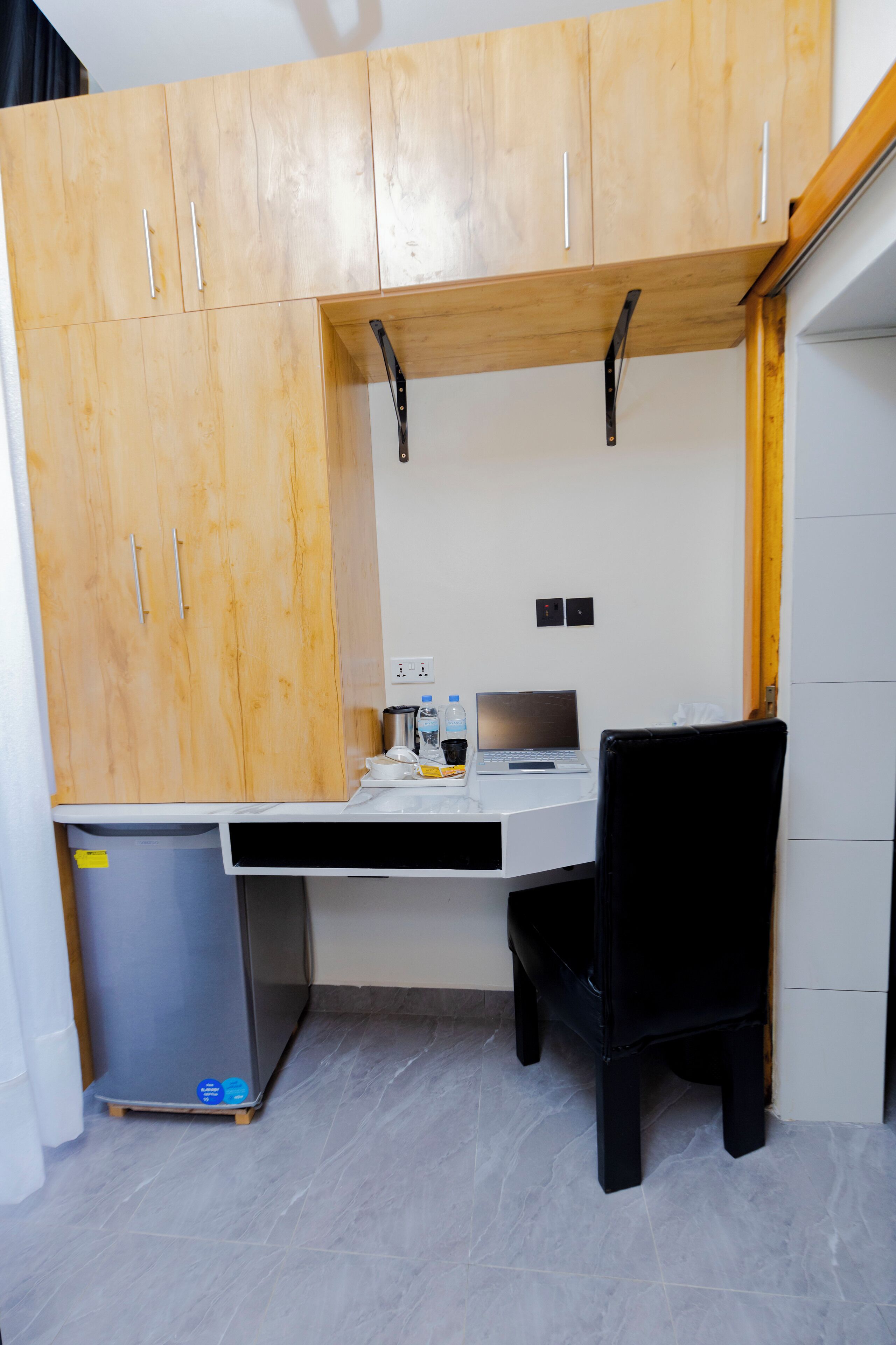 A compact office space with a wooden desk, a chair, a small refrigerator, and a laptop.