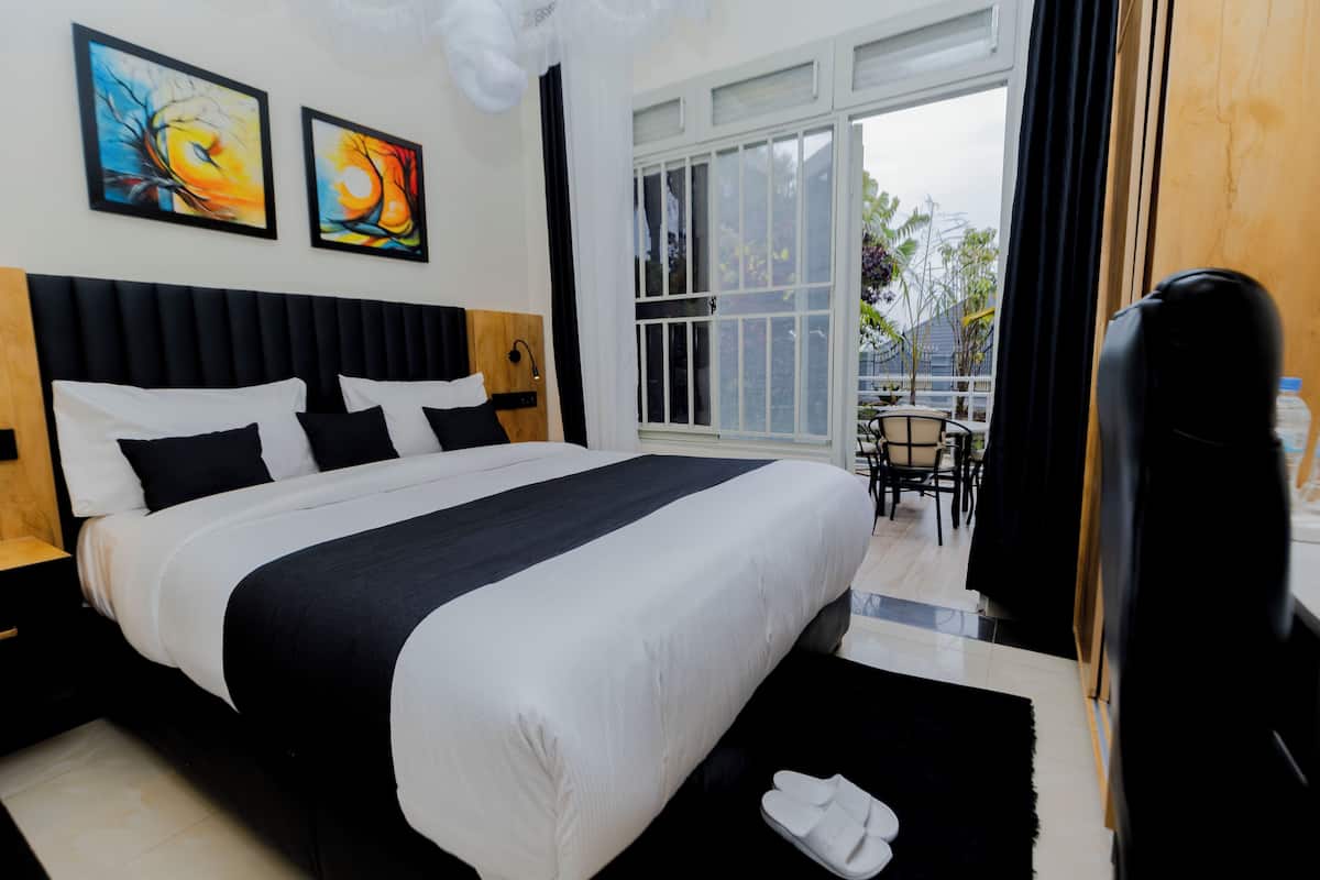 Quarto de hotel com uma cama grande, cabeceira preta, duas pinturas na parede, janela com vista para o exterior e uma pequena mesa com cadeiras.