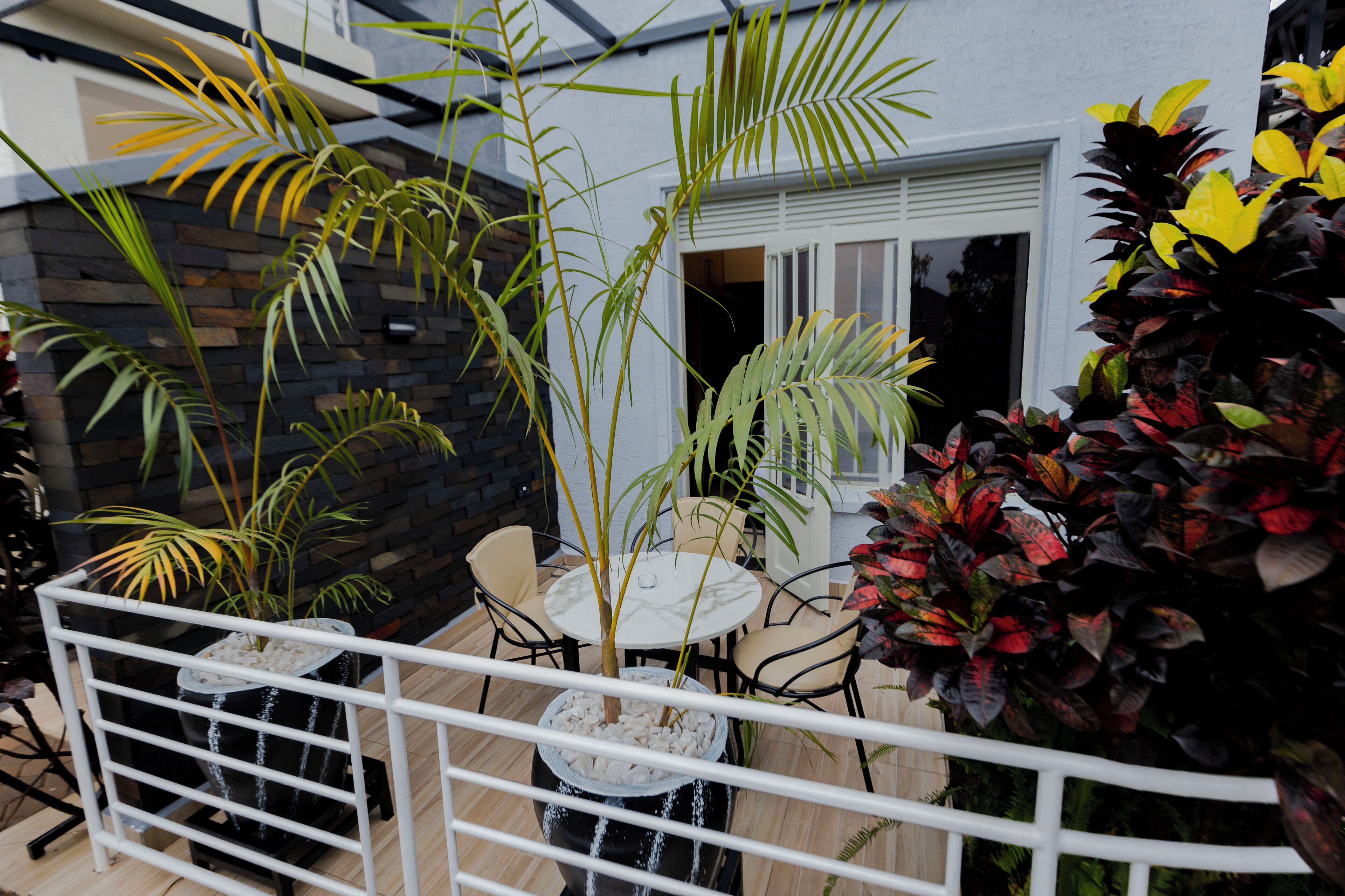 Un balcon avec une balustrade blanche, une petite table et des chaises. On y trouve différentes plantes en pot et un élément décoratif avec de l’eau.