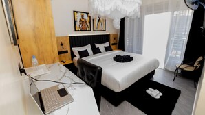 A modern hotel room with a bed, a desk with a laptop, a chair, a bedside table, a bedside lamp, a window with curtains, and a wall-mounted artwork.