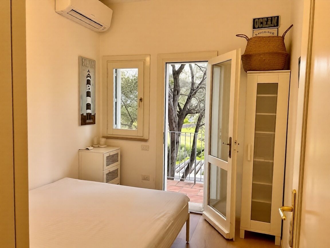 A bedroom with a bed, a dresser, a window, a door leading to a balcony, and a wall-mounted air conditioner.