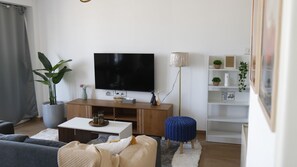 Ein Wohnzimmer mit Flachbildfernseher, einer Holzunterhaltungsecke, Topfpflanzen, einem Couchtisch, einem blauen Pouf und einem weißen Regal mit Dekoration.